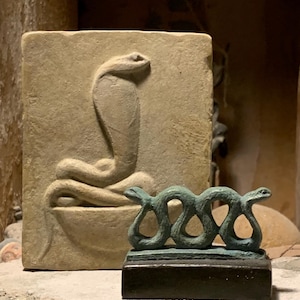 Puede incluir: Una placa rectangular de piedra beige con un relieve de una cobra enroscada en un cuenco, situada detrás de una pequeña escultura sobre base oscura de dos serpientes entrelazadas de color verde azulado con superficies texturizadas.
