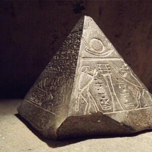 May include: A small, grey stone pyramid with hieroglyphics carved into its sides. The pyramid is approximately 15 cm tall and has a square base.