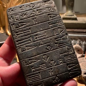 Puede incluir: Una tableta rectangular de color gris oscuro con escritura cuneiforme. El antiguo artefacto tiene filas de marcas y símbolos en forma de cuña. La tableta está sostenida en la mano de una persona, con otros objetos antiguos en el fondo.