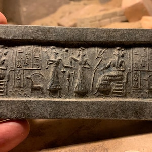 May include: A rectangular gray stone seal featuring intricate relief carvings of figures, animals, and cuneiform-style inscriptions. The surface displays a horizontal scene with standing and seated individuals, a small animal, and vertical columns of etched symbols.