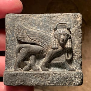 Puede incluir: Una placa cuadrada de piedra gris oscuro con un relieve de una esfinge alada. La criatura tiene cabeza humana con un tocado tradicional, cuerpo de león de perfil y grandes alas detalladas que se extienden hacia arriba sobre la superficie.