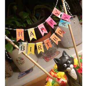 May include: A colorful 'Happy Birthday' banner with watercolor-style flags hangs above a black cat cake topper. The cake topper has white whiskers and large eyes.