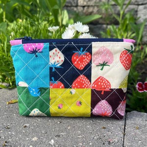 May include: A quilted pouch with a patchwork design featuring strawberries on various colored squares. The pouch has a navy blue zipper and a pink trim. The background is blurred with greenery.