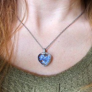 Könnte beinhalten: Eine silberne Kettenkette mit einem herzförmigen Anhänger. Der Anhänger ist mit getrockneten blauen Vergissmeinnicht-Blüten gefüllt.