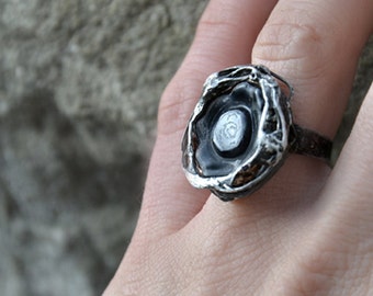 May include: A silver ring with a black and white stone set in a textured, organic-shaped bezel. The ring is worn on a person's finger.