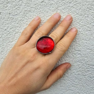 Puede incluir: Un anillo grande y redondo con una vibrante piedra roja translúcida engastada en una banda de metal plateado. El anillo se muestra en una mano sobre un fondo gris claro texturizado. La piedra parece tener variaciones internas.