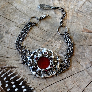 Puede incluir: Un brazalete de cadena de plata con un cabujón de vidrio rojo engastado en un marco de filigrana de plata. El marco está decorado con pequeños diamantes de imitación rojos.