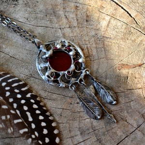 Puede incluir: Un collar de plata con un colgante redondo que presenta un cabujón de vidrio rojo rodeado de filigrana de plata. El colgante tiene dos plumas de plata que cuelgan de él.