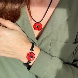 May include: A black cord necklace and bracelet with red poppy flower charms. The necklace and bracelet are worn by a person.