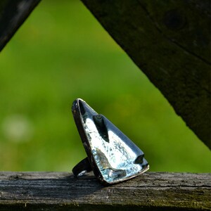Puede incluir: Un anillo triangular de vidrio con una banda plateada. El anillo está sobre una superficie de madera.