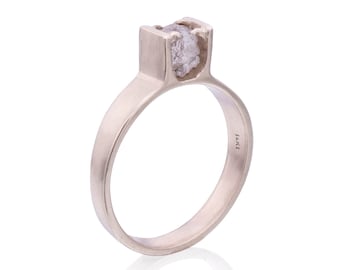 Könnte beinhalten: Ein silberner Ring mit einem rohen, klaren Diamanten, der in einer quadratischen Fassung sitzt. Der Ring ist innen mit "14K" gekennzeichnet.
