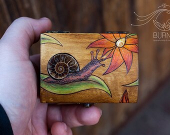 Puede incluir: Una caja de madera con un diseño pintado a mano de un caracol y una flor. El caracol es marrón y la flor es naranja y amarilla. La caja se sostiene en una mano y tiene el texto "BURNEX".