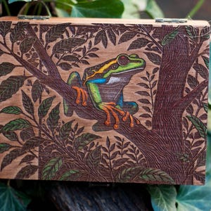 Può includere: Una scatola di legno con una scultura dettagliata di una rana verde e arancione seduta su un ramo con foglie. La scatola è in legno chiaro e ha una finitura naturale.