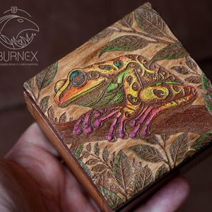Può includere: Una scatola di legno con una scultura colorata e dettagliata di una rana su un ramo frondoso. La rana è verde, gialla e rosa con macchie nere. La scatola è un'ottima aggiunta a qualsiasi arredamento per la casa.