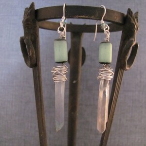 Puede incluir: Pendientes hechos a mano con una cuenta cilíndrica verde claro, alambre de plata y una punta de cristal transparente. Los pendientes cuelgan de ganchos de plata. Se exhiben en un soporte de metal decorativo.
