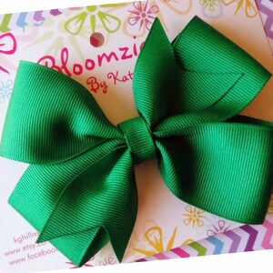 May include: A large green bow made from grosgrain ribbon. The bow is tied with a center loop and is ready to be attached to a hair clip or headband. The bow is on a white background with a floral design and the text "Bloomzie By Kat" in pink.