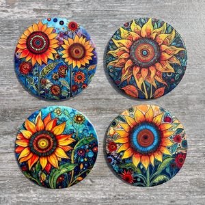 May include: Four round coasters with vibrant sunflower designs. Each coaster features a unique sunflower illustration with yellow, orange, and red petals, set against a blue or dark background. The coasters are a decorative accessory.