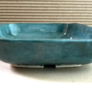 May include: A handmade, rectangular ceramic dish with a teal glaze. The dish has a slightly irregular shape and sits on two small, white feet. The interior of the dish has a glossy finish, and the exterior has a matte appearance.