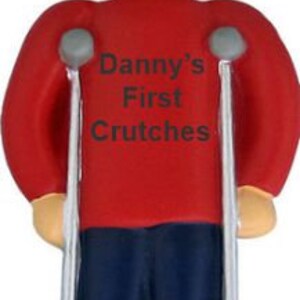 May include: A figurine of a person with a cast on one leg, wearing a red shirt and a green hat. The shirt has the text "Danny's First Crutches". The figurine is holding crutches.