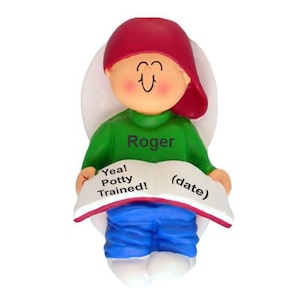 May include: A figurine of a person wearing a red cap, green shirt with the name "Roger", and blue pants. The figurine holds a book that reads "Yea! Potty Trained!" and "(date)". The figurine has a white background.