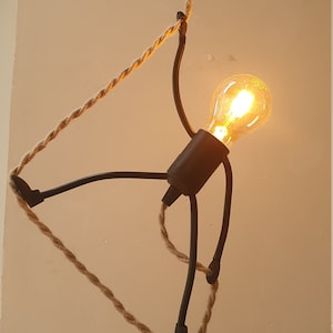 Pode incluir: Um candeeiro de metal preto em forma de figura de pau que segura uma lâmpada. A lâmpada está acesa e o candeeiro está suspenso do teto por uma corda grossa e natural.