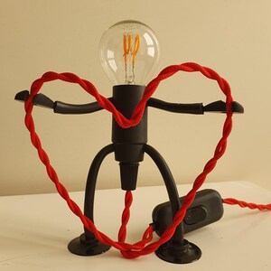 May include: A black table lamp shaped like a person with a red cord wrapped around its arms, forming a heart shape. The lamp has a clear light bulb with a filament.