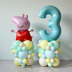 May include: Two balloon centerpieces for a birthday celebration. One features a pink pig balloon wearing a red dress, the other a light blue number 3 balloon. Both are atop columns of pastel-colored balloons.
