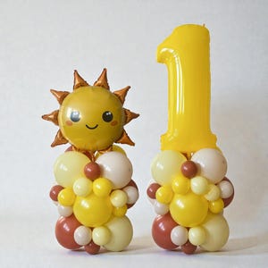 May include: Two balloon decorations for a first birthday. One is a smiling sun with brown rays. The other is a large yellow number one balloon. Both are on balloon bases with yellow, white, and brown balloons.