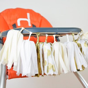 Puede incluir: Una guirnalda de borlas de papel de seda blanco y dorado cuelga del tablero de una trona. Las borlas est&aacute;n hechas de papel de seda blanco con lunares dorados y papel de seda dorado.