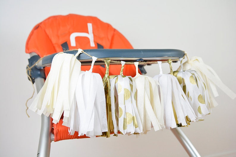 Puede incluir: Una guirnalda de borlas de papel de seda blanco y dorado cuelga del tablero de una trona. Las borlas est&aacute;n hechas de papel de seda blanco con lunares dorados y papel de seda dorado.