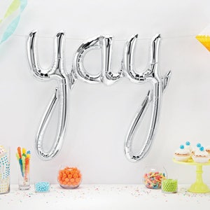 May include: Silver foil balloon spelling "yay" with colorful decorations and treats on a white table.