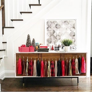 May include: A wooden console table with a black metal base, decorated with a garland of red, black, and white tassel streamers. The table is styled with a gray and white abstract painting, a small potted plant, and a stack of books.