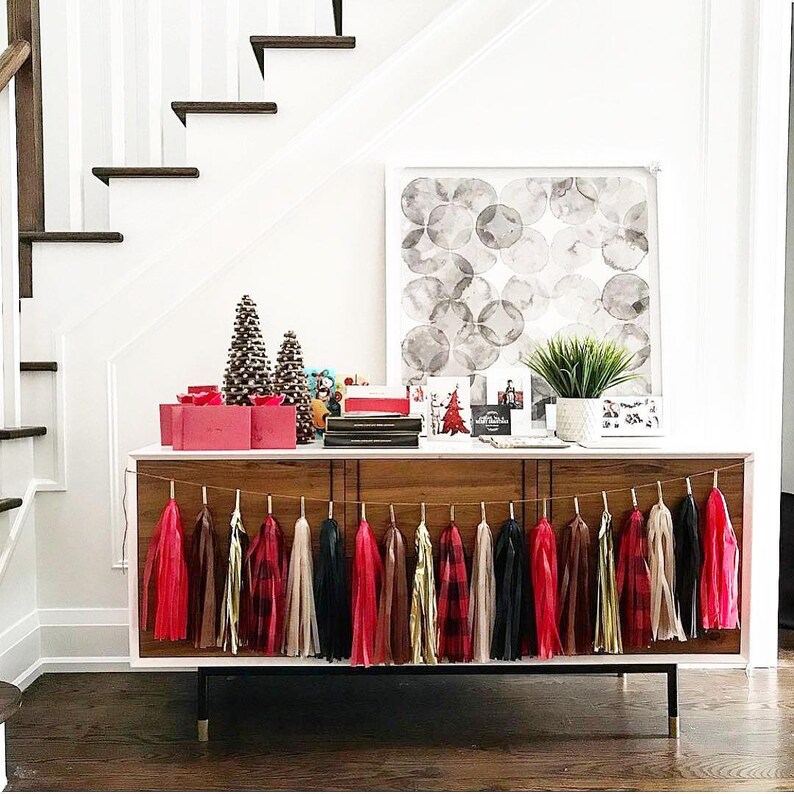 May include: A wooden console table with a black metal base, decorated with a garland of red, black, and white tassel streamers. The table is styled with a gray and white abstract painting, a small potted plant, and a stack of books.