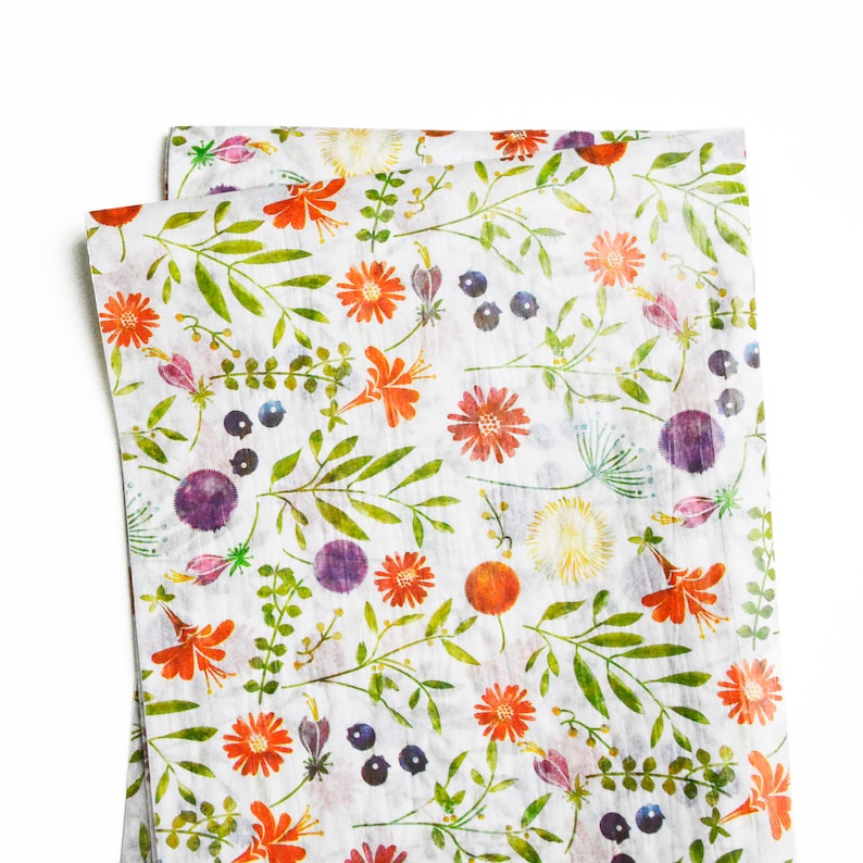Op de afbeelding: Wit inpakpapier met een bloemenpatroon met oranje, rode, paarse en gele bloemen, groene bladeren en kleine blauwe bessen.