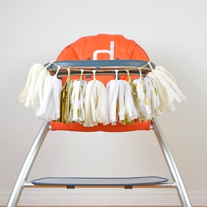 Puede incluir: Una trona con una guirnalda de borlas blancas y doradas drapeada en la parte superior. La silla es naranja y plateada.