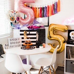 May include: A white table with two white chairs, a black and white striped gift bag, a gold number five balloon, a pink number five balloon, a skull balloon with floral details, a sign that says "Spooky Boo Day Paisley", and cupcakes on a cake stand.