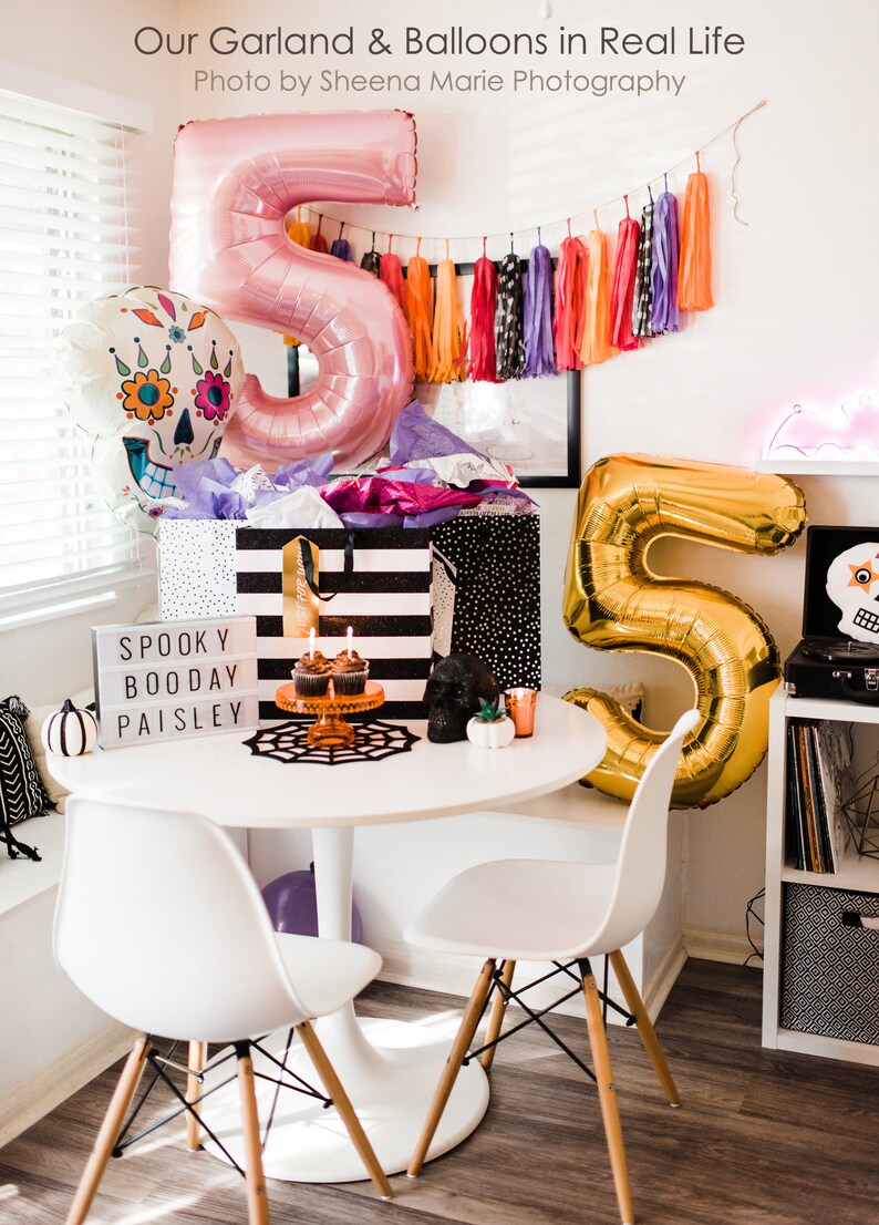 May include: A white table with two white chairs, a black and white striped gift bag, a gold number five balloon, a pink number five balloon, a skull balloon with floral details, a sign that says "Spooky Boo Day Paisley", and cupcakes on a cake stand.