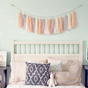 May include: A white bed with a white headboard and a peach and white tassel garland above it. The bed has a white, peach, and gray patterned pillow and a white unicorn stuffed animal. There are two nightstands with lamps and books on them.