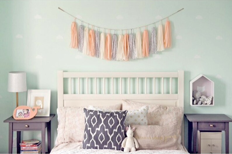 May include: A white bed with a white headboard and a peach and white tassel garland above it. The bed has a white, peach, and gray patterned pillow and a white unicorn stuffed animal. There are two nightstands with lamps and books on them.
