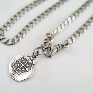 May include: A silver chain necklace with a silver coin pendant featuring a cross design. The chain is a curb style and the pendant is attached to the chain with a lobster clasp.