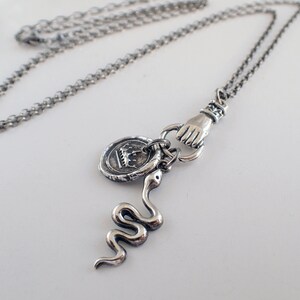 May include: A silver chain necklace with a silver hand charm, a silver snake charm, and a silver coin charm with a crown design.