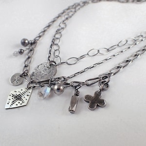 May include: A silver necklace with multiple chains and charms, including a diamond-shaped charm with a geometric pattern, a round charm with a textured surface, a small cross charm, and a long, thin charm with a textured surface.
