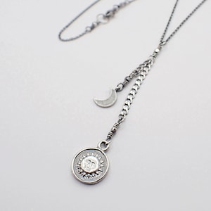 May include: A silver-toned necklace featuring a sun pendant and a crescent moon charm. The sun pendant has a detailed face design within a circular frame. The necklace has a delicate chain and a unique Y-shaped design, perfect for celestial-themed jewelry.