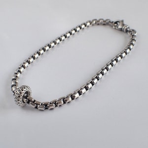 May include: A silver chain bracelet with a decorative round charm. The chain is made of small, linked squares.