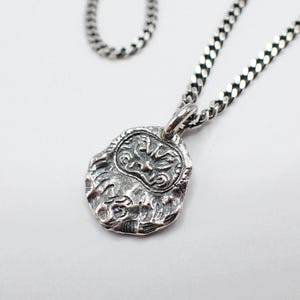 May include: A silver-toned pendant necklace featuring a textured, oval-shaped pendant with a detailed face design. The pendant hangs from a curb chain, creating a classic and detailed accessory. The necklace is displayed against a white background.