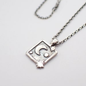 May include: Silver-toned pendant necklace featuring a diamond-shaped pendant with a crescent moon, stars, and a dot design. The pendant hangs from a delicate chain. The necklace is set against a white background.