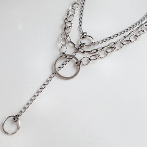 May include: A silver layered chain necklace with a large circle pendant. The necklace has three different chain styles.