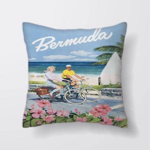 Puede incluir: Una almohada de estilo póster de viaje vintage con una pareja montando bicicletas en un camino con una playa y palmeras de fondo. El texto "Bermuda" está escrito en una fuente blanca cursiva en la parte superior de la almohada.