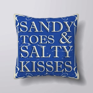 Puede incluir: Cojín cuadrado azul con las palabras "SANDY TOES & SALTY KISSES" en blanco, con letras de estilo vintage. El cojín tiene un aspecto envejecido, con un borde blanco y un fondo claro.