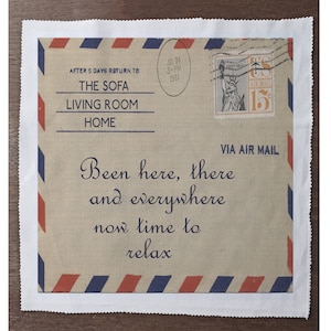 May include: A white fabric coaster with a vintage-style airmail envelope design. The envelope has red and blue stripes, a stamp with a picture of the Statue of Liberty, and the text "AFTER 5 DAYS RETURN TO THE SOFA LIVING ROOM HOME" and "Been here, there and everywhere now time to relax".